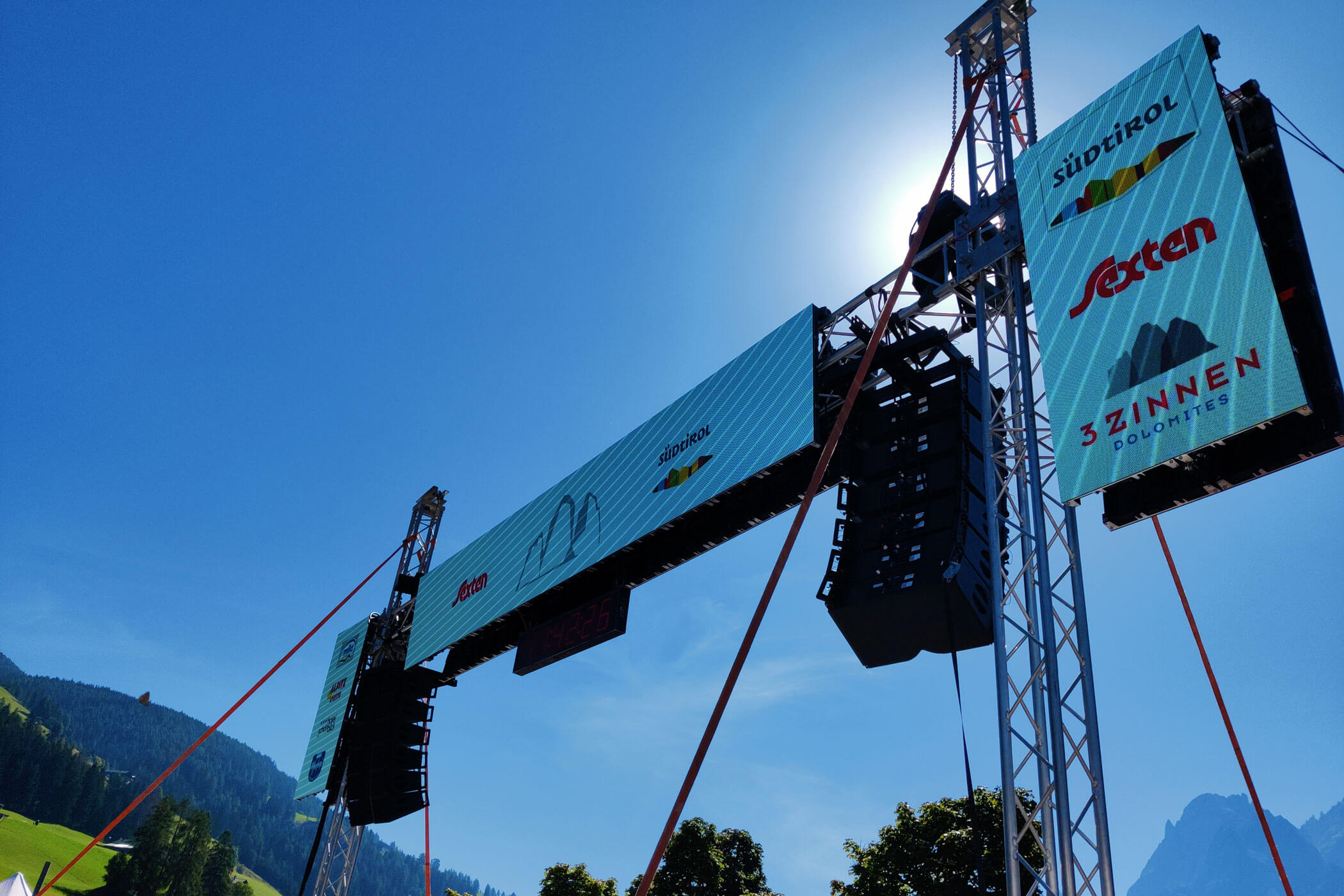 Eine Konzertbühne im Freien mit großen Lautsprechern und Bannern vor dem Hintergrund eines strahlend blauen Himmels und einer Berglandschaft. - HJPA Pro