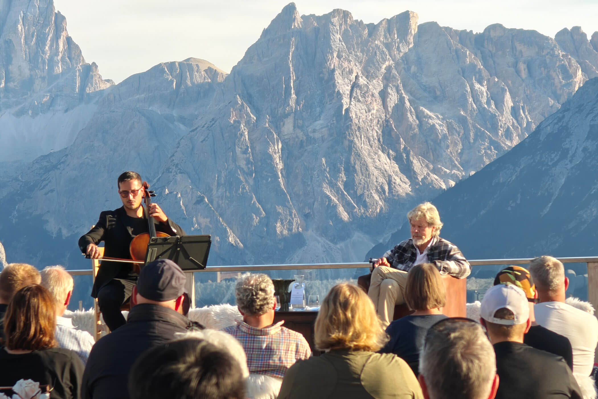 Ein Cellist und ein Redner treten im Freien vor Publikum auf, im Hintergrund sind Berge zu sehen. - HJPA Pro