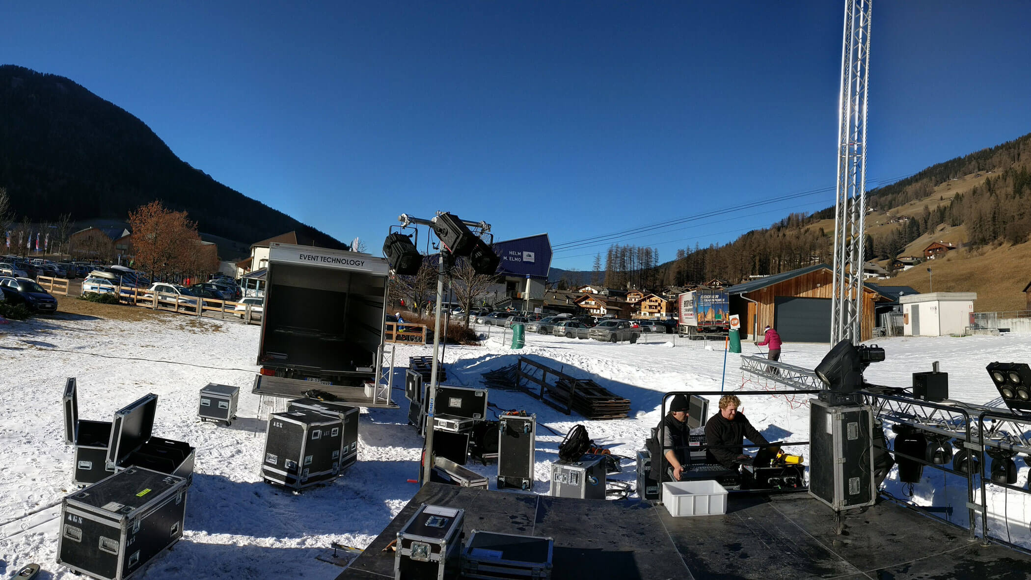 Aufbau einer Freiluftbühne mit Ausrüstungskoffern, zwei Arbeitern, Schnee auf dem Boden und Bergen im Hintergrund. - HJPA Pro