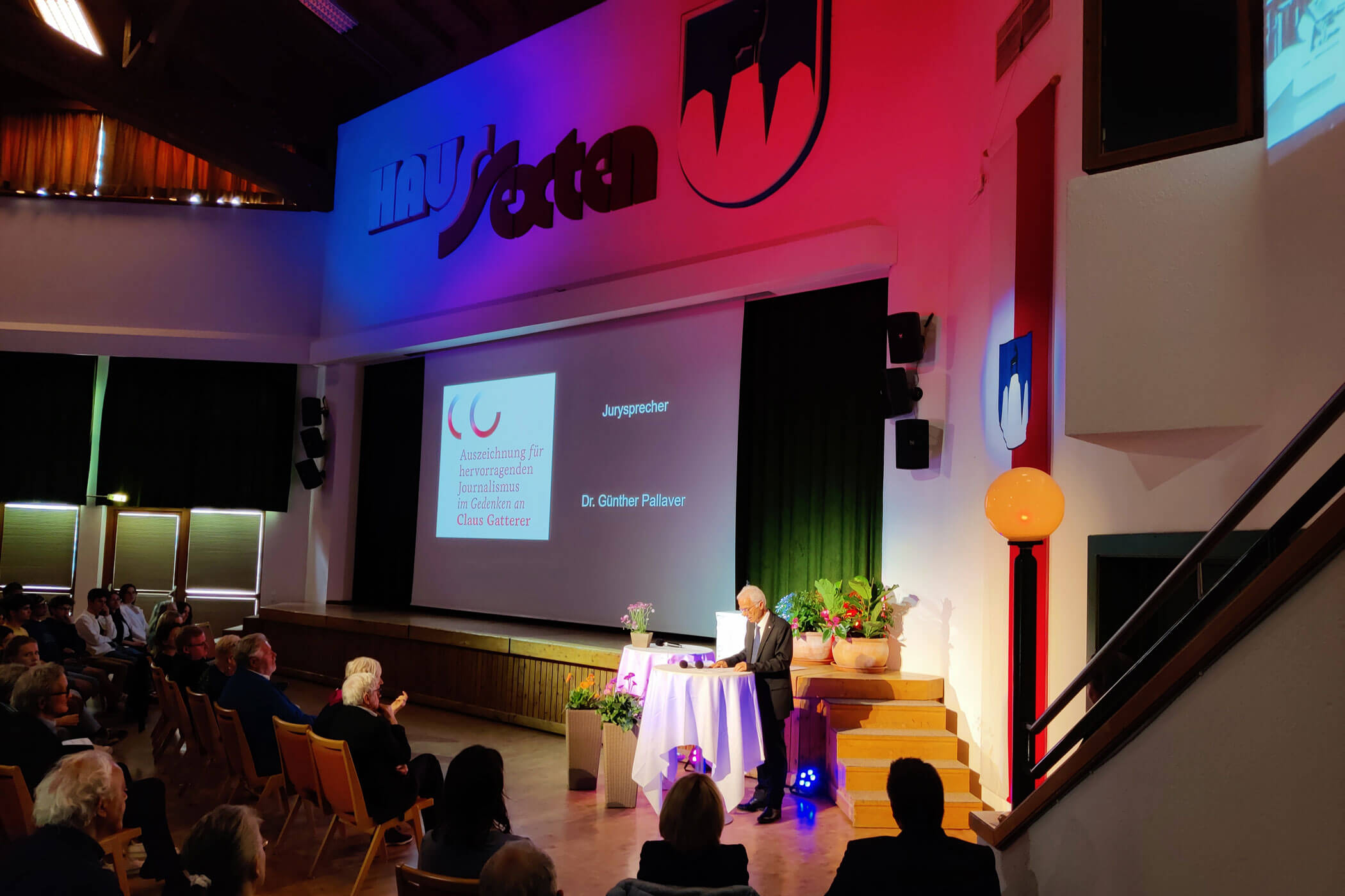 Ein Redner hält auf einem Podium in einem farbenfrohen, modernen Saal eine Rede, während das Publikum aufmerksam zuhört. - HJPA Pro
