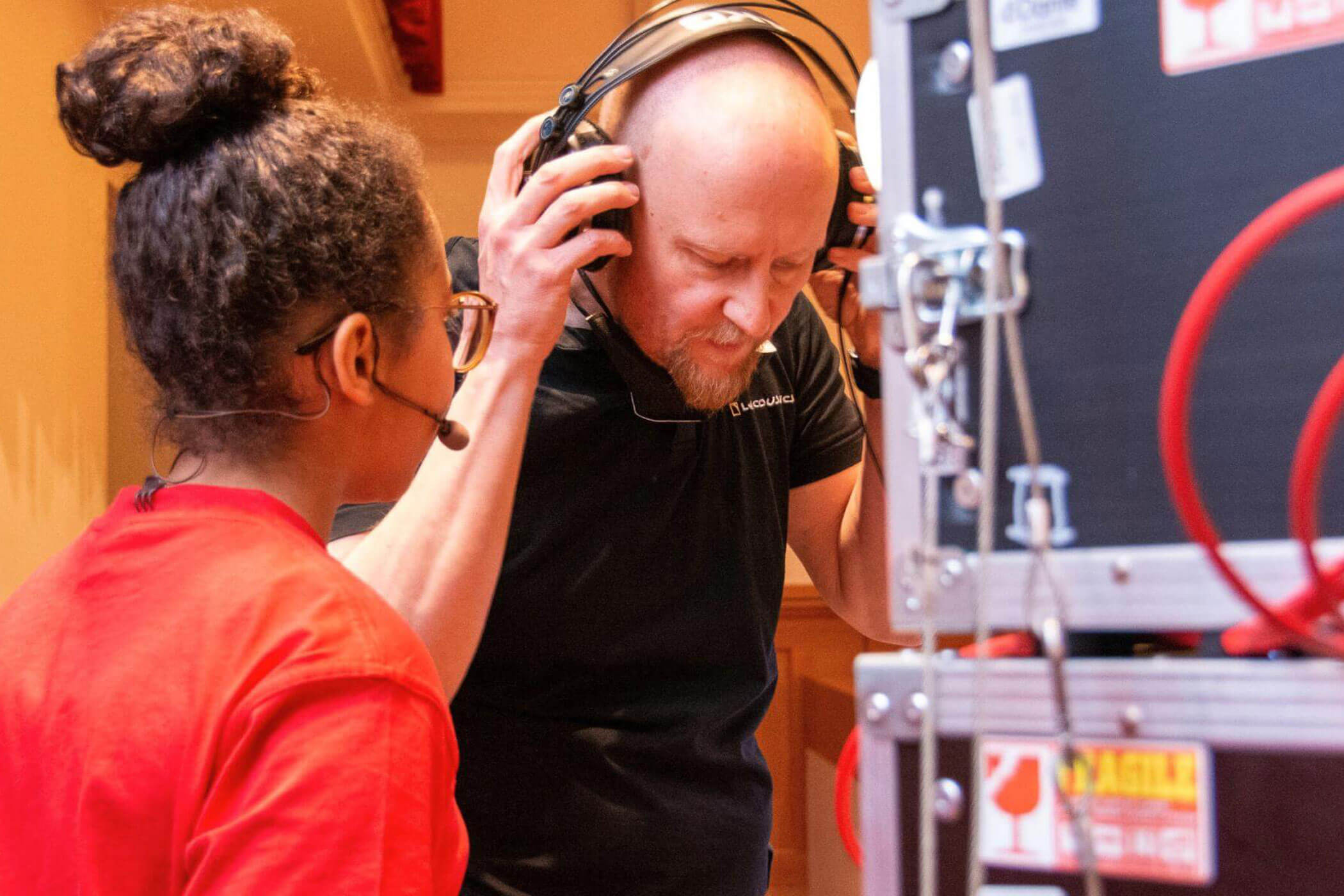 Zwei Personen hinter der Bühne: Eine trägt ein Headset, die andere stellt in der Nähe der technischen Anlage ihre Kopfhörer ein. - HJPA Pro