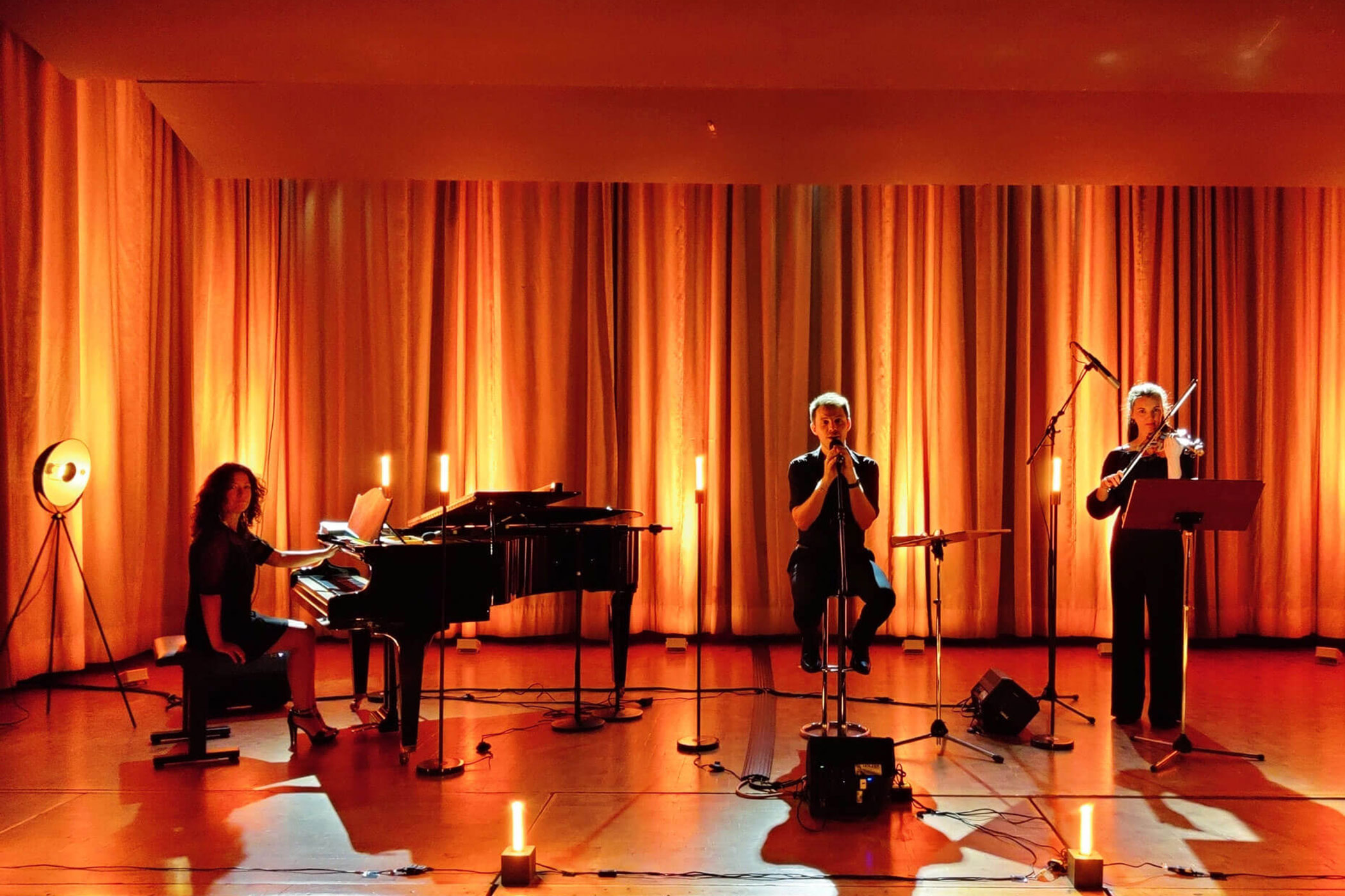 Drei Musiker treten auf der Bühne mit Klavier, Gesang und Violine auf, beleuchtet von warmem orangefarbenem und gelbem Licht. - HJPA Pro