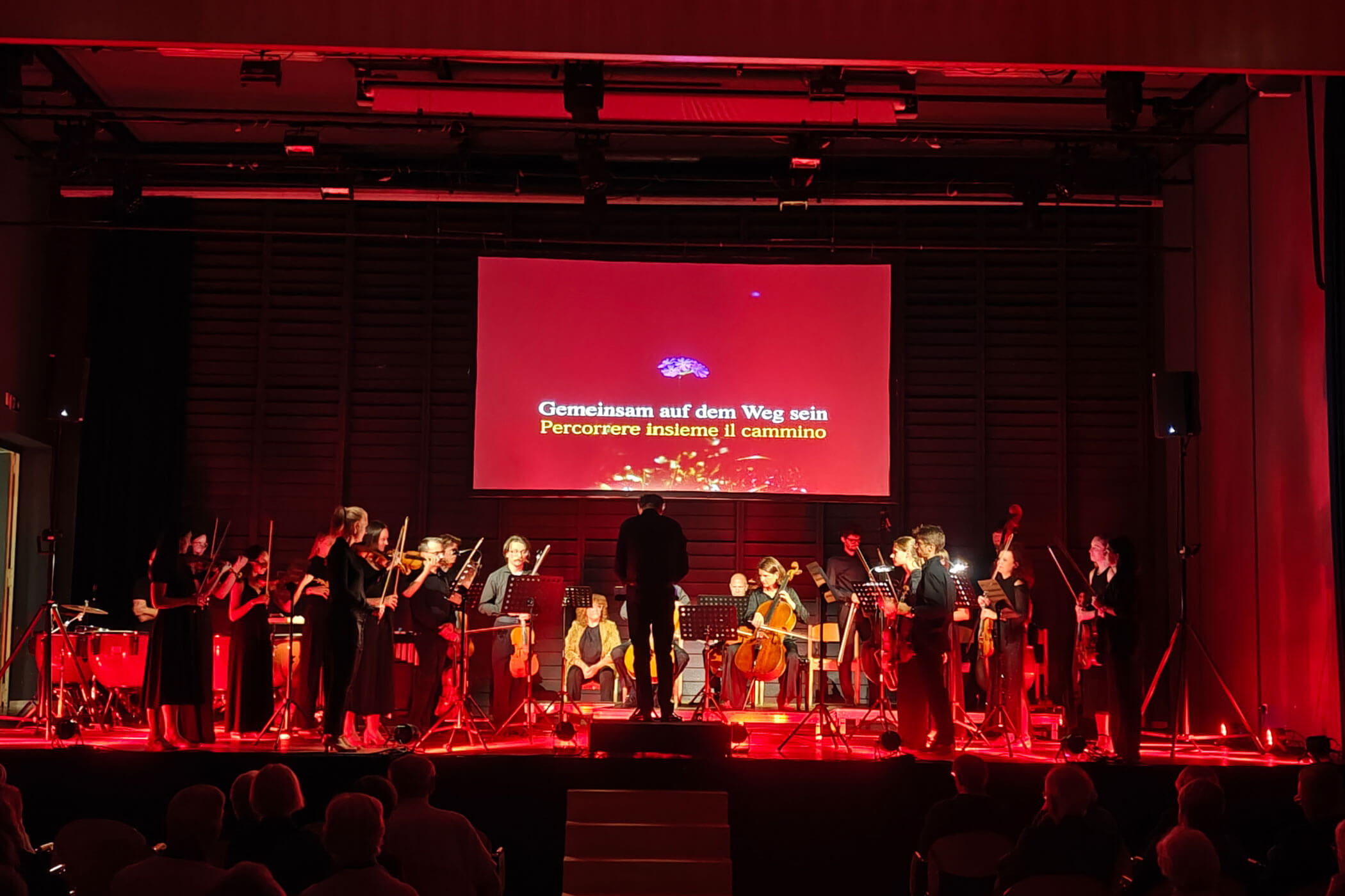 Ein Orchester spielt auf der Bühne bei rotem Licht; hinter den Musikern wird Text auf Deutsch und Italienisch auf eine Leinwand projiziert. - HJPA Pro